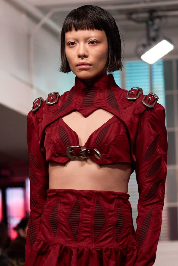 Fashion Portrait, model wearing sculptural crimson co-ord set with cutout panels and silver buckle hardware, runway lighting