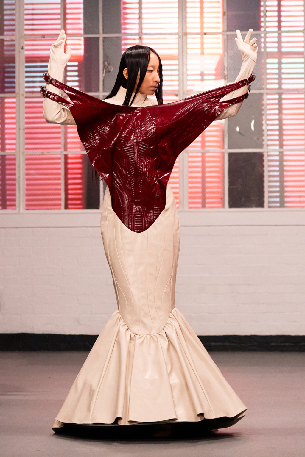Fashion show, LFW AW26 404: Safety Not Found, model in sculptural cream mermaid gown with dramatic crimson patent leather bodice, arms raised, runway lighting