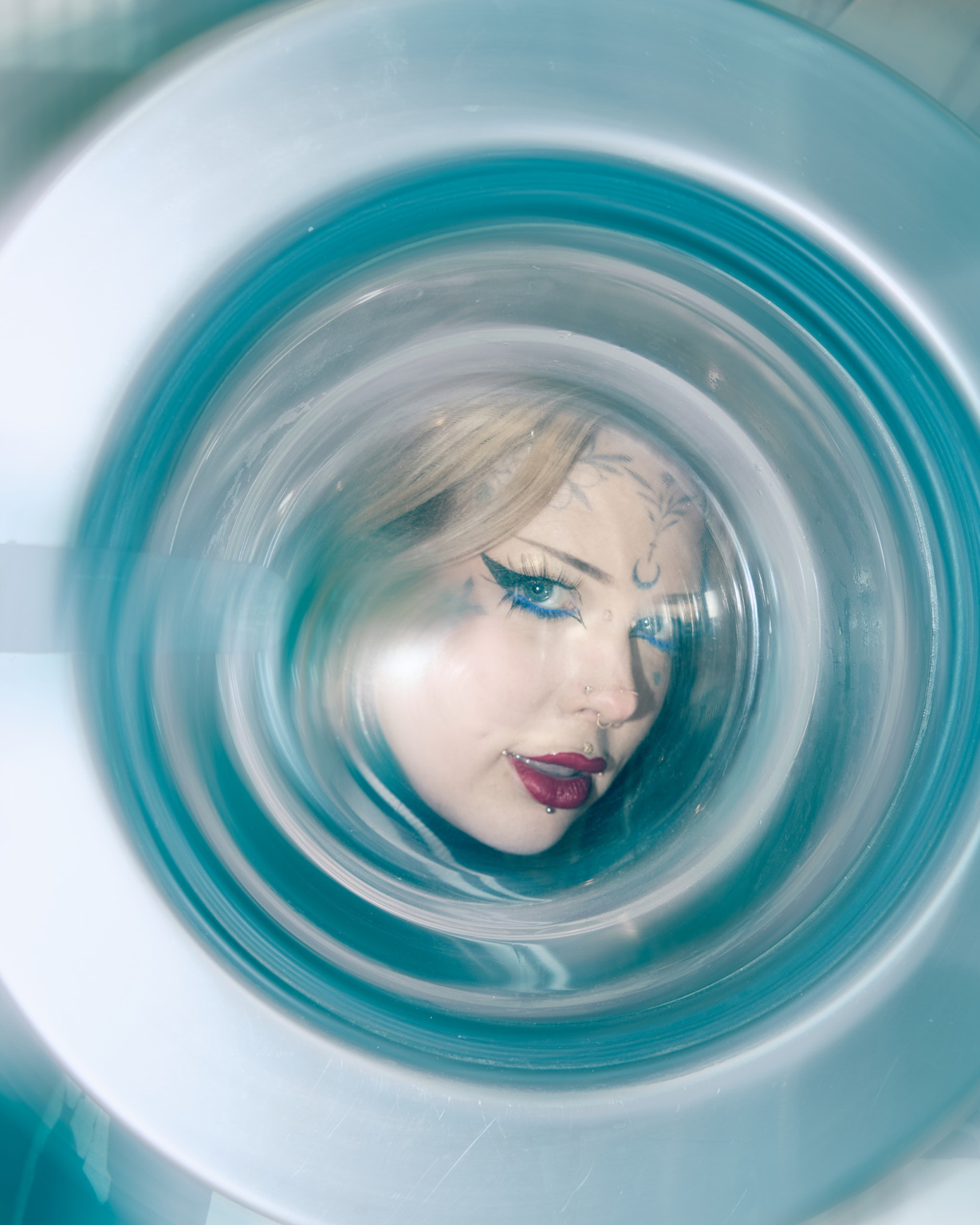 Editorial portrait, model's face framed behind washing machine door, harsh flash lighting