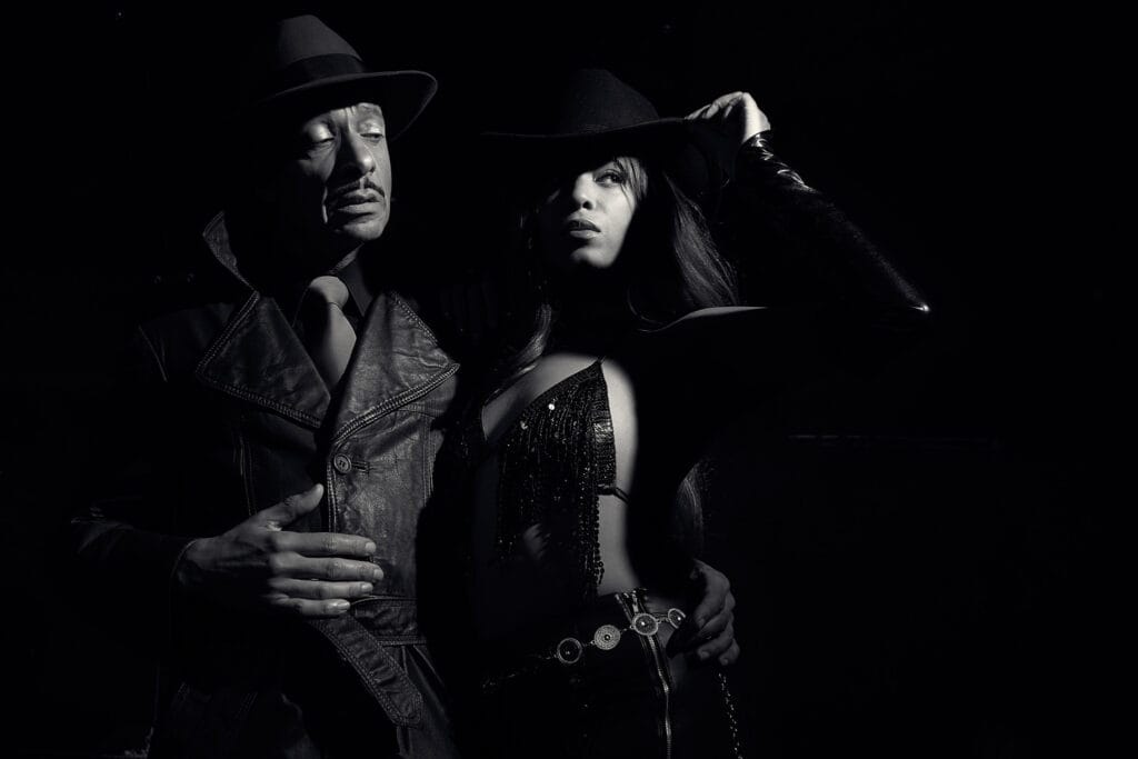Fashion noir portrait, Sin City inspired, two models both wearing hats, male model in leather trench coat and tie, female model in sparkling fringed top and leather trousers with metal belt, dramatic noir lighting