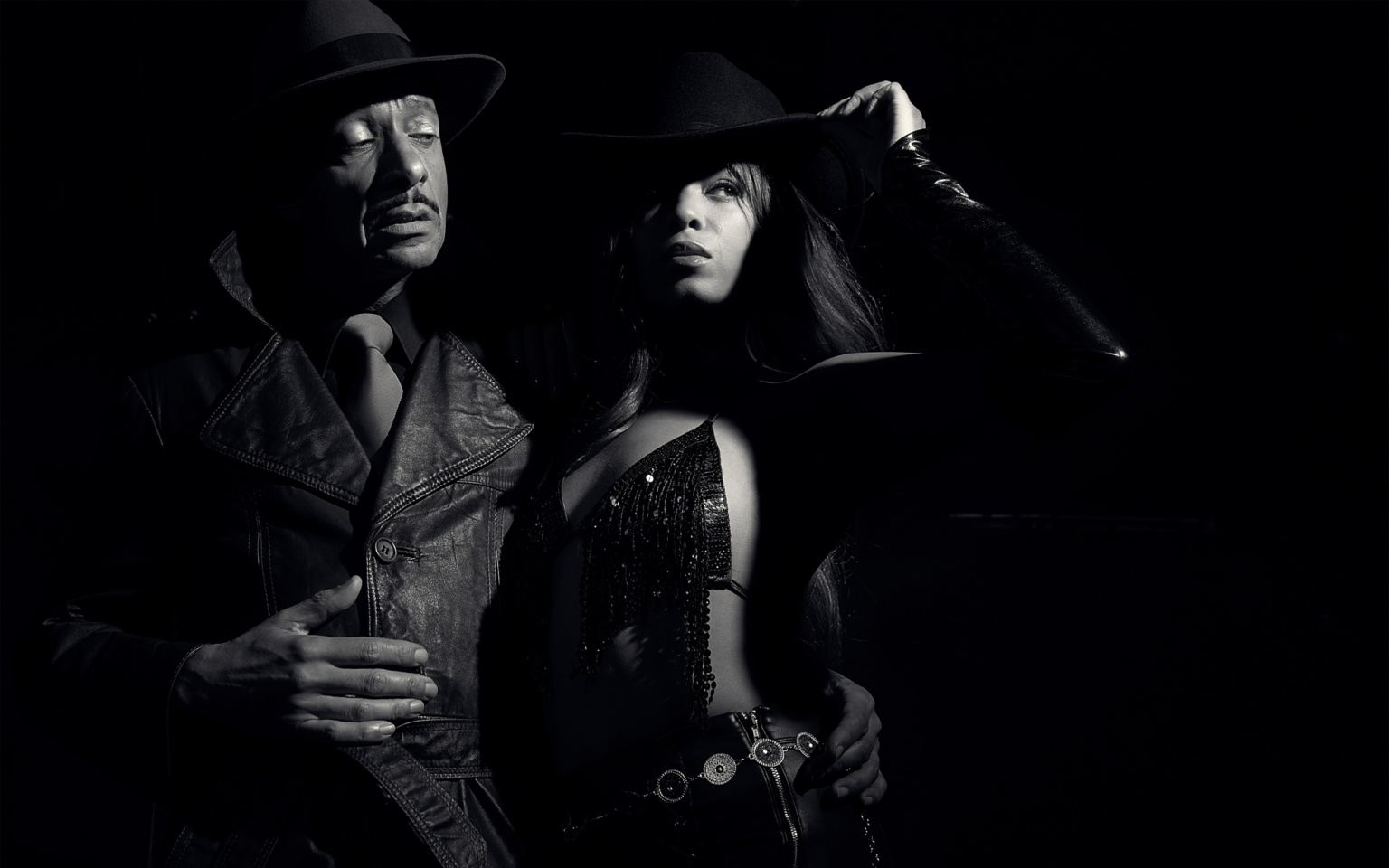 Fashion noir portrait, Sin City inspired, two models both wearing hats, male model in leather trench coat and tie, female model in sparkling fringed top and leather trousers with metal belt, dramatic noir lighting